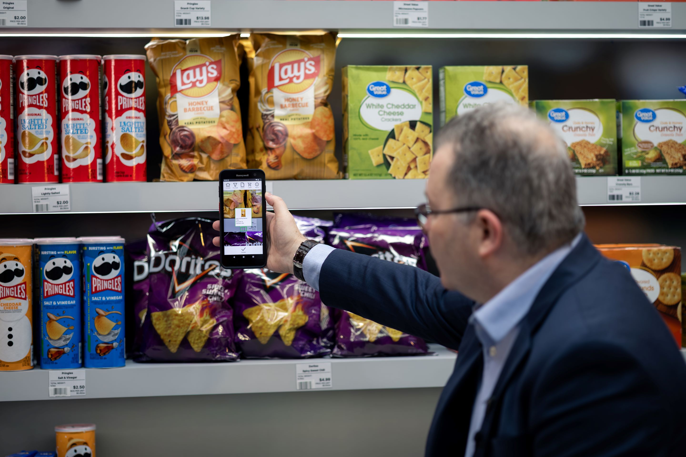 A person holds a smartphone while browsing a grocery store, comparing products on the shelves filled with various packaged items.